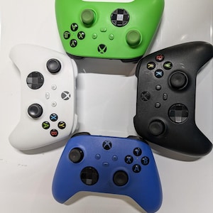 Peut inclure: Quatre manettes de jeu vidéo sans fil disposées en cercle. La collection comprend une manette vert vif, une manette noir mat, une manette blanche et une manette bleue, chacune équipée de deux joysticks et de boutons de commande.
