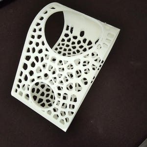 Könnte beinhalten: Weißes 3D-gedrucktes Objekt mit geometrischem Muster und zwei kreisförmigen Öffnungen. Das Objekt ist gebogen und hat ein gitterartiges Design.