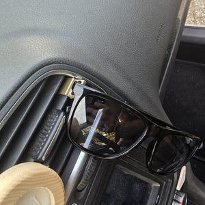 Può includere: Occhiali da sole neri con un clip dorato attaccato alla bocchetta di ventilazione di un'auto. Gli occhiali da sole sono appoggiati sul cruscotto dell'auto.