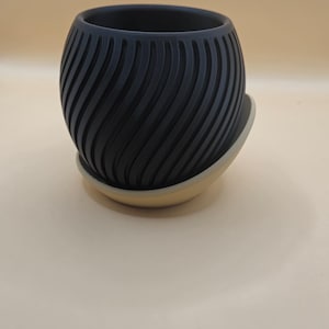 Könnte beinhalten: Ein schwarzer Keramiktopf mit einem wellenförmigen, strukturierten Design. Der Topf hat eine passende beige Untertasse.