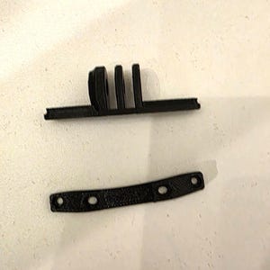 Könnte beinhalten: Zwei schwarze Kamera-Zubehörteile aus Kunststoff. Ein Teil hat einen gebogenen Abschnitt mit drei vertikalen Schlitzen und einer flachen Basis. Das andere ist ein schwarzer Riemen mit drei Löchern.