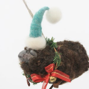 May include: A brown, fuzzy Christmas ornament shaped like an animal, wearing a teal and white Santa hat. It has a red bow with gold trim and a small bell. The ornament is decorated with greenery.