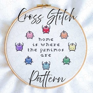 May include: A cross-stitch pattern in a wooden hoop. The design features colorful pixelated creatures surrounding the text "home is where the junimos are." The words "Cross Stitch" and "Pattern" are written in cursive.
