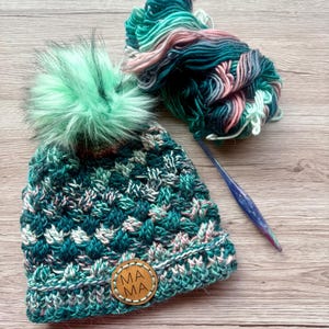 May include: A green, pink, and white crocheted hat with a pom-pom and a brown leather patch that says "mama".