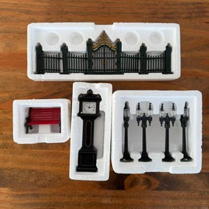 May include: A collection of miniature village accessories packaged in white styrofoam, including a black metal gate with gold accents, a red park bench, a black grandfather clock with a white face, and four black street lamps with clear tops.