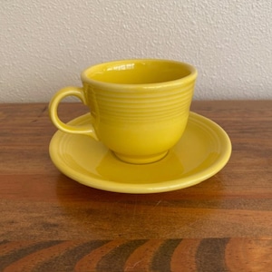 Könnte beinhalten: Ein leuchtend gelbes Set aus Teetasse und passender Untertasse aus Keramik. Die Teetasse verfügt über einen abgerundeten Henkel und eine Reihe horizontaler, konzentrischer Ringe, die außen am Körper eingeprägt sind. Die Untertasse ist glatt und rund.