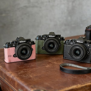 May include: Three Nikon Zf cameras with different colored leather cases: pink, green, and black. The cameras are on a brown surface. The pink camera has the name "Kate" on it. The green camera has the letter "H" on it.
