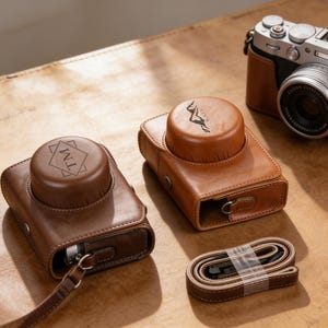 May include: A collection of brown leather camera cases and a camera with a silver lens. One case has a monogram, another has a graphic. A matching brown strap is also included. The items are on a brown surface.