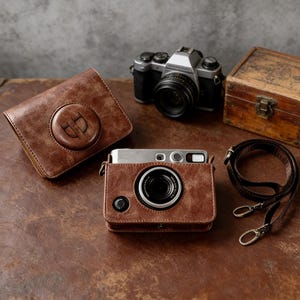 Könnte beinhalten: Ein braunes Leder-Kameraetui und -Gurt-Set mit einer silbernen Kamera. Das Etui hat ein kreisförmiges geprägtes Design. Eine Vintage-Holzkiste und eine weitere Kamera befinden sich ebenfalls im Bild.