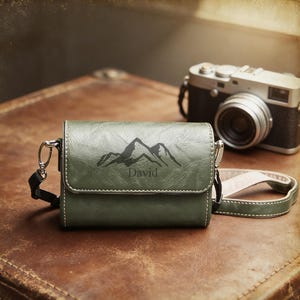 Könnte beinhalten: Eine olivgrüne Kameratasche aus Leder mit einem Bergmotiv und dem Namen "David" auf der Vorderseite. Die Tasche hat einen Schultergurt und einen silbernen Verschluss. Eine Vintage-Kamera befindet sich im Hintergrund.