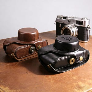 May include: Two leather camera cases, one brown and one black, with a classic camera in the background. The brown case has a lighter brown stitching and brass-colored hardware. The black case has the initials "LR" on the top.