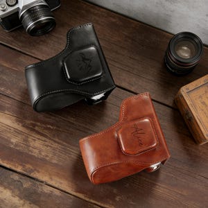May include: Two leather camera cases, one black and one brown, are displayed on a wooden surface. The black case has white stitching and a mountain logo with the text "ZAN". The brown case has the name "Alice" on it.