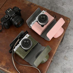 May include: A black digital camera with a lens and two other cameras in leather cases. One case is pink, and the other is olive green. The cases are open, revealing the camera lenses.