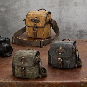 Può includere: Tre borse per fotocamera in tela nei toni del beige, verde oliva e grigio antracite. Ogni borsa presenta una chiusura con patta in pelle con fibbia, finiture in pelle e una tracolla. La borsa beige ha il nome "Chiara D" sulla tracolla.