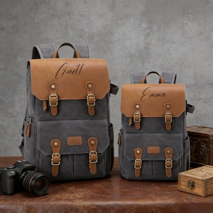 Peut inclure: Deux sacs à dos en toile grise avec des détails en cuir marron. Le plus grand porte le nom "Anett" et le plus petit "Emma" sur le rabat en cuir. Les deux ont plusieurs poches et des sangles réglables, avec des boucles métalliques.