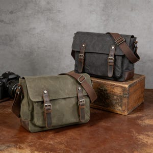 Peut inclure: Deux sacs messagers en toile, vert olive et gris anthracite, avec des détails en cuir et des sangles réglables. Les sacs ont une forme rectangulaire avec une fermeture à rabat. Ils sont conçus pour transporter des objets et sont présentés sur une surface en bois.