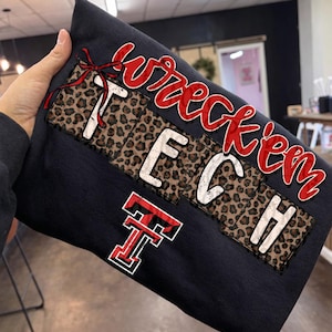 Puede incluir: Una sudadera azul marino con las palabras "Wreck'em TECH" en letras rojas y blancas. La palabra "TECH" está en un parche con estampado de leopardo. También es visible un logotipo "T" rojo y blanco.