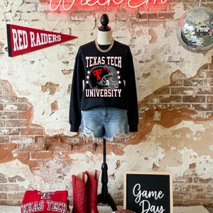 May include: A black Texas Tech University sweatshirt with a football helmet graphic, paired with denim shorts, red cowboy boots, and a football. A red pennant reads "Red Raiders". A sign says "Game Day".