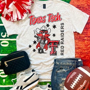 May include: A light gray Texas Tech Red Raiders t-shirt with a cartoon mascot graphic, paired with denim shorts, sneakers, a football, and a black bag. A red and white pom-pom is also included.
