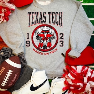 May include: A gray Texas Tech sweatshirt with a graphic of a cartoon character holding guns, the words "TEXAS TECH", "1923", and "WRECK'EM TECH". The image also includes a football, high-top sneakers, a red baseball cap, and red and white pom-poms.