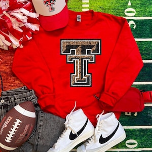 May include: A red sweatshirt with a large letter "T" design, a red and white baseball cap, and white high-top trainers with black swooshes. A football and denim jeans are also in the image. The sweatshirt features a leopard print pattern within the letter.