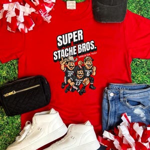 May include: A red t-shirt with the text "SUPER STACHE BROS." and a graphic of three cartoon football players. The image also includes white high-top sneakers, a black crossbody bag, a black trucker hat, blue jeans, and red and white pom-poms.
