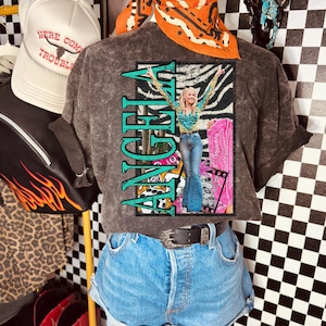 Könnte beinhalten: Präsentation eines grauen T-Shirts mit dem Namen "ANGELA" in türkisfarbenen Buchstaben und einer Grafik einer Frau im Country-Western-Stil. Die Präsentation umfasst Jeansshorts, einen Gürtel, ein Bandana und eine Trucker-Mütze mit dem Text "HERE COME TROUBLE".