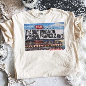 May include: Off-white t-shirt with a graphic of Levi's Stadium and the message "THE ONLY THING MORE POWERFUL THAN HATE IS LOVE." The design includes a stadium scene and the words "Seguimos Aqui."