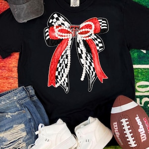 May include: Black t-shirt with a large bow design featuring red and black checkered patterns, a pearl strand, and the letter "T" in red. The image includes white high-top sneakers, a football, a denim cap, and jeans.
