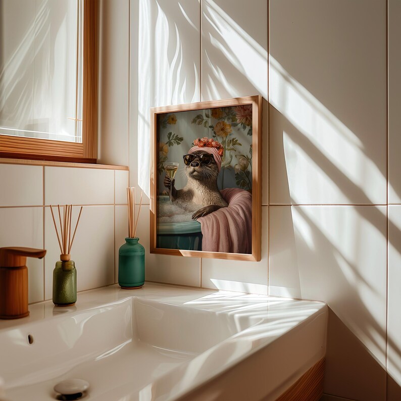 May include: A framed print featuring an otter in a bathtub, wearing sunglasses and a headscarf, holding a champagne glass. The artwork has a vintage style with floral wallpaper in the background. The frame is made of light-colored wood.