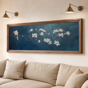 Puede incluir: Una obra de arte rectangular enmarcada que presenta una rama de flores de magnolia blancas sobre un fondo azul oscuro. El marco de madera rodea la imagen, y la obra de arte está montada en una pared de color claro. Dos apliques de latón están montados encima de la obra de arte.