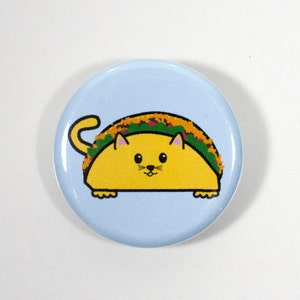 Puede incluir: Un bot&oacute;n azul con un gato de dibujos animados que lleva una c&aacute;scara de taco amarilla. El gato tiene orejas rosas y una nariz negra.