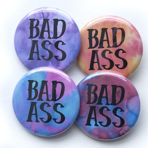 Może przedstawiać: Cztery okrągłe guziki z czarnym napisem "BAD ASS" na tle w stylu tie-dye w odcieniach fioletu, niebieskiego, różu i pomarańczy. Guziki są błyszczące i mają agrafkę z tyłu.