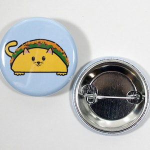 Puede incluir: Un bot&oacute;n azul con un gato de dibujos animados que lleva un taco. El gato es amarillo con la nariz rosa y bigotes. El taco est&aacute; relleno de ingredientes verdes y marrones. La parte posterior del bot&oacute;n es plateada con un alfiler.