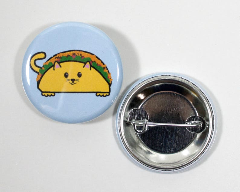 Puede incluir: Un bot&oacute;n azul con un gato de dibujos animados que lleva un taco. El gato es amarillo con la nariz rosa y bigotes. El taco est&aacute; relleno de ingredientes verdes y marrones. La parte posterior del bot&oacute;n es plateada con un alfiler.