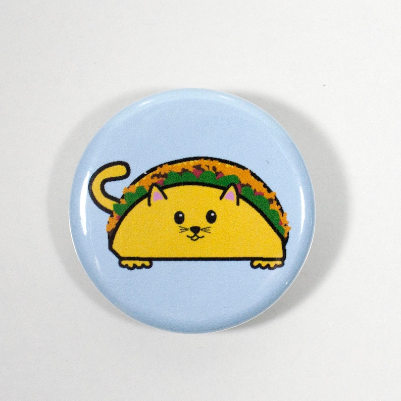 Puede incluir: Un bot&oacute;n azul con un gato de dibujos animados que lleva una c&aacute;scara de taco amarilla. El gato tiene orejas rosas y una nariz negra.