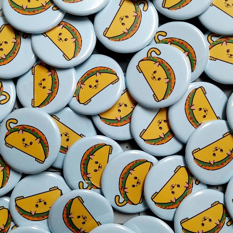 Puede incluir: Una colecci&oacute;n de botones redondos de color azul claro con un contorno blanco. Cada bot&oacute;n presenta un gato de dibujos animados con un disfraz de taco. Los gatos est&aacute;n en varias poses, algunos est&aacute;n sentados, otros est&aacute;n de pie y otros est&aacute;n acostados. Los tacos est&aacute;n rellenos de diferentes ingredientes, como queso, lechuga y tomates.