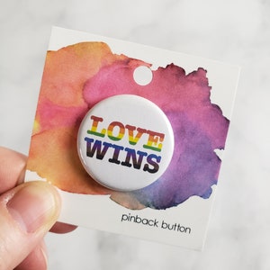 May include: A white pinback button with the text "LOVE WINS" in rainbow colours. The button is on a white card with a watercolour background.