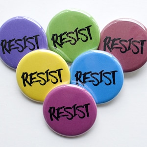 Może przedstawiać: Sześć okrągłych guzików z napisem "RESIST" w kolorze czarnym na każdym. Guziki są w różnych kolorach: fioletowym, zielonym, bordowym, żółtym, niebieskim i różowym.