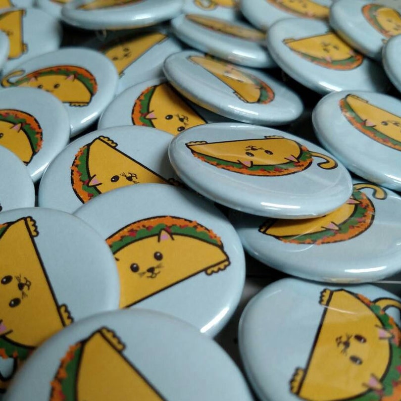 Puede incluir: Una colecci&oacute;n de botones redondos azules y blancos con un gato de dibujos animados que lleva un disfraz de taco. El gato es amarillo con una cara blanca y ojos negros. El taco es marr&oacute;n con lechuga verde y relleno rojo.