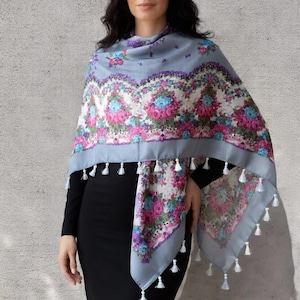 May include: A woman wearing a gray shawl with a floral pattern in pink, blue, and purple. The shawl has white tassels along the edges. The shawl is 100cm x 100cm.