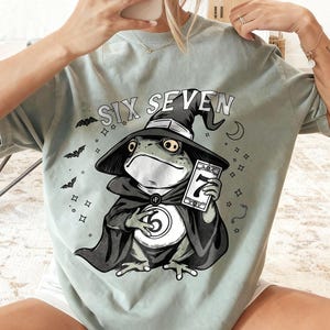 May include: Sage green t-shirt featuring a cartoon frog dressed as a wizard, holding a card with the number seven. The frog wears a black hat and cape, with the words "SIX SEVEN" above. Bats and stars surround the frog.