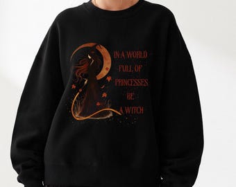 May include: Black sweatshirt with the text "IN A WORLD FULL OF PRINCESSES BE A WITCH" in orange. The design features a witch silhouette with a crescent moon and autumn leaves. The sweatshirt has a crew neck and long sleeves.