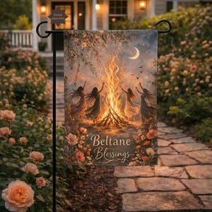 Puede incluir: Una bandera de jardín de doble cara que representa una escena de hoguera con figuras bailando alrededor de un fuego bajo una luna creciente. La bandera tiene las palabras "Beltane Blessings" y está rodeada de flores.