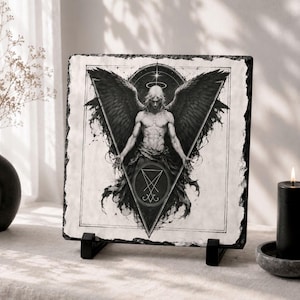 May include: A black and white illustration of an angel with large wings, a halo, and a triangle design. The artwork is displayed on a square slate tile, propped up on a black stand. A black candle and vase are also in the image.
