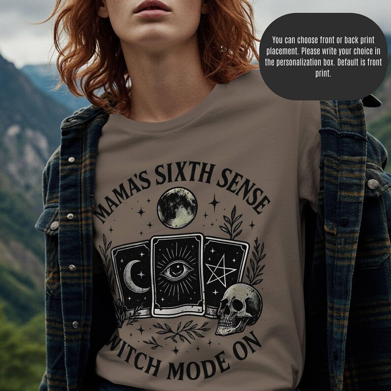 K&ouml;nnte beinhalten: Ein taupefarbenes T-Shirt mit einer Grafik mit Tarotkarten, einem Totenkopf und einem Mond. Der Text lautet "MAMA'S SIXTH SENSE WITCH MODE ON". Das Shirt wird von einer Person mit roten Haaren und einer karierten Jacke getragen. Das Bild enth&auml;lt auch Text &uuml;ber die Platzierung des Drucks.