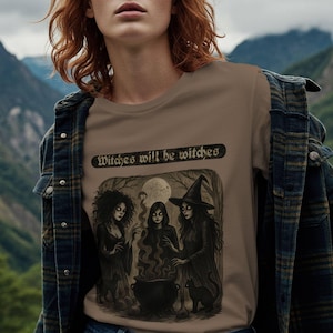 Pode incluir: Camiseta bege com uma ilustração escura de três bruxas em volta de um caldeirão, com um gato preto. O texto "Witches will be witches" está acima da ilustração. O design é colocado em um fundo de lua cheia e árvores.