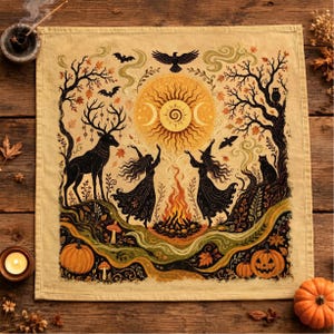 May include: A square beige fabric cloth featuring a detailed illustration of two figures dancing around a campfire. The scene includes a large sun with moon phases, a stag, bats, a raven, an owl, a black cat, pumpkins, and autumn trees.