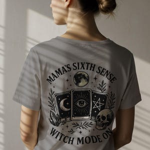 K&ouml;nnte beinhalten: Hellgraues T-Shirt mit einem grafischen Design mit Tarotkarten, einem Totenkopf und dem Text "MAMA'S SIXTH SENSE WITCH MODE ON". Das Design umfasst einen Halbmond, Sterne und botanische Elemente.