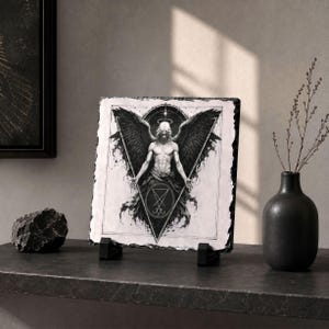 Puede incluir: Ilustraci&oacute;n en blanco y negro de una figura alada con halo, enmarcada en un tri&aacute;ngulo y un c&iacute;rculo. La obra de arte se muestra sobre una piedra cuadrada texturizada. Se apoya en dos soportes negros, con un jarr&oacute;n negro y una piedra en la superficie.